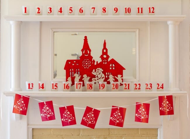 A festive advent calendar with numbers 1 to 24 placed over a fireplace, featuring a mirror decorated with a red silhouette of santa claus and a village, and red envelopes with white botanical designs hanging below.