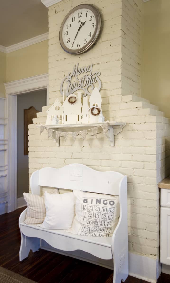 A cozy corner featuring a white bench with a cushion under a decorative mantel decorated with a "merry christmas" sign, small houses, and a large circular wall clock on a white brick pillar.