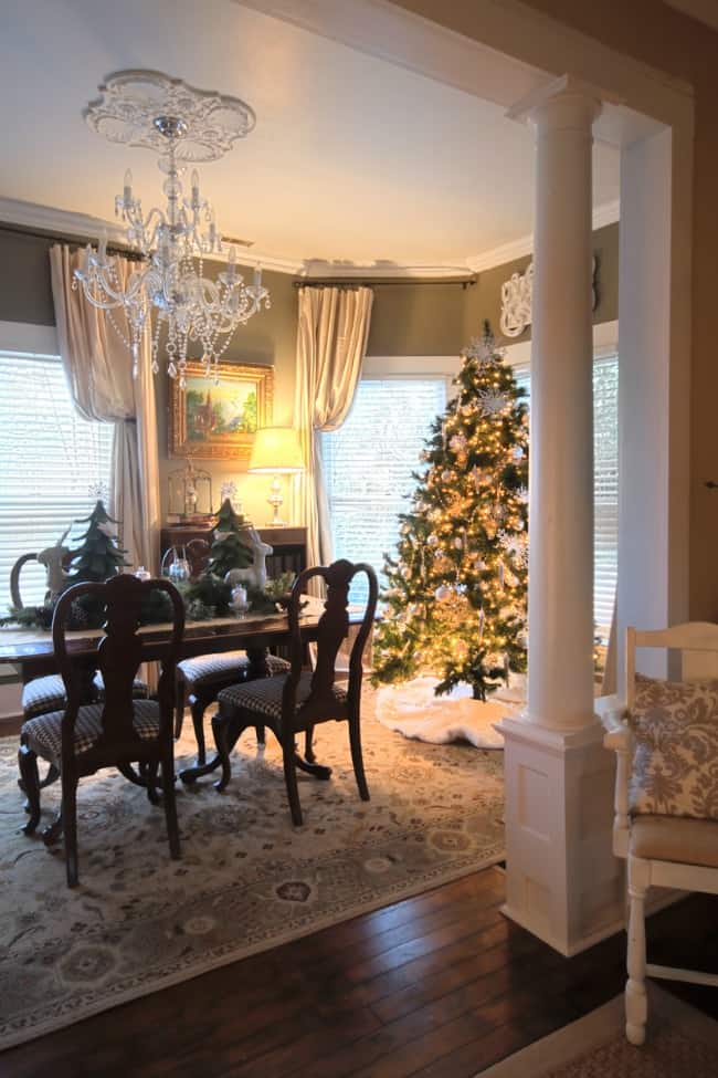 Elegant dining room decorated for christmas, featuring a lit christmas tree in the corner, a chandelier above, and a table set with greenery. white chair and cream curtains complement the warm ambiance.