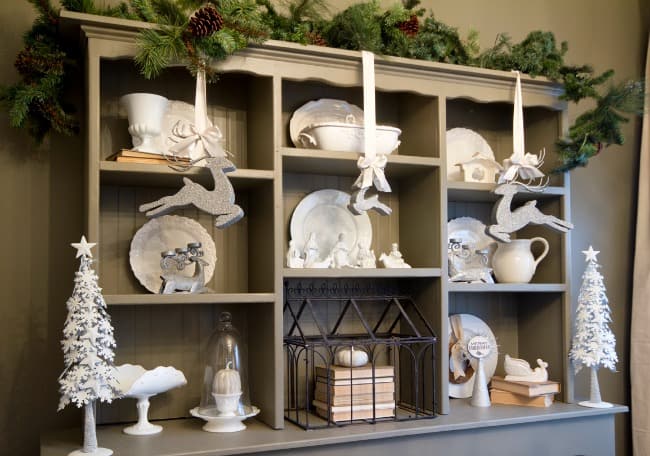 A festive holiday-themed shelf display featuring white decorative items including reindeer, elephants, trees, and assorted figurines, accented with green garlands above.