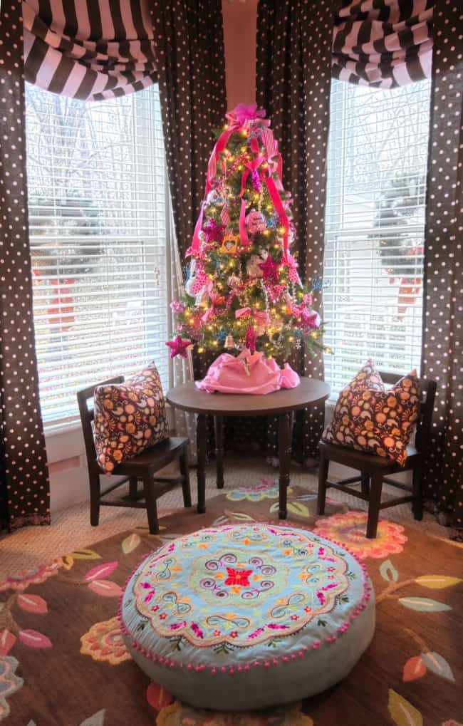 A vibrant and festive room featuring a brightly decorated Christmas tree with pink and brown accents, flanked by chairs with patterned cushions, set against a window with striped curtains.