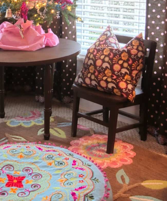 A cozy room corner featuring a dark wooden chair with a pink patterned cushion, a small round table with a vibrant pink decoration, a colorful floral rug, and a lit Christmas tree in the background.