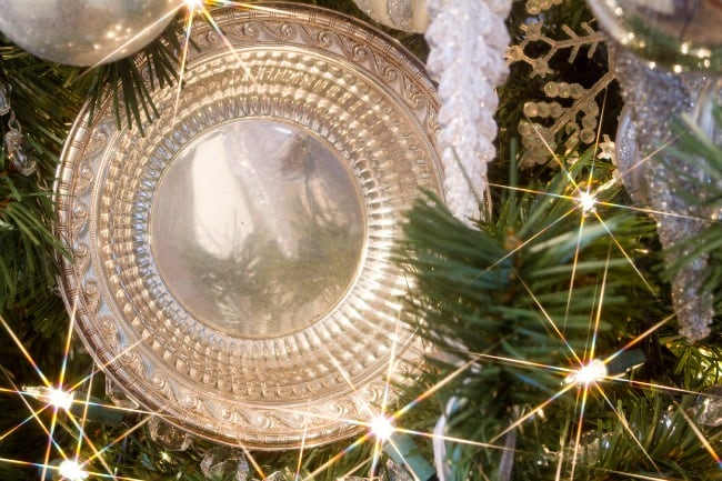 A close-up view of a shiny, ornate silver christmas ornament reflecting light, nestled among green pine branches with twinkling white lights.