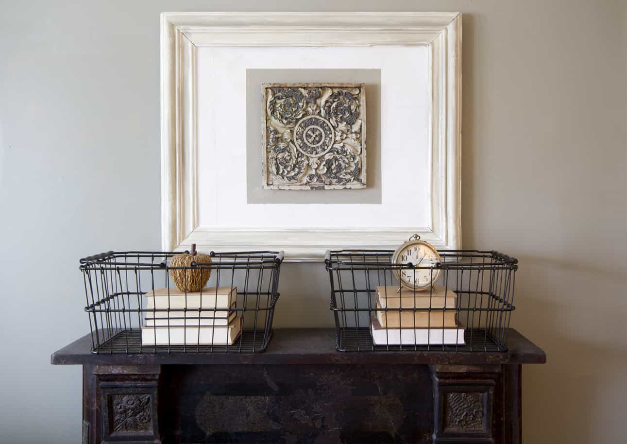 Minimalist home decor with symmetrically arranged metal baskets containing books and decorative items on a wooden console table, set against a cream wall with an ornate framed artwork.
