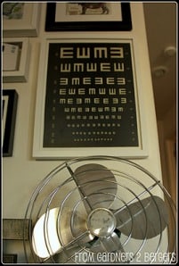 Vintage-style oscillating fan in the foreground with a collection of framed eye chart posters hanging on the wall in the background.