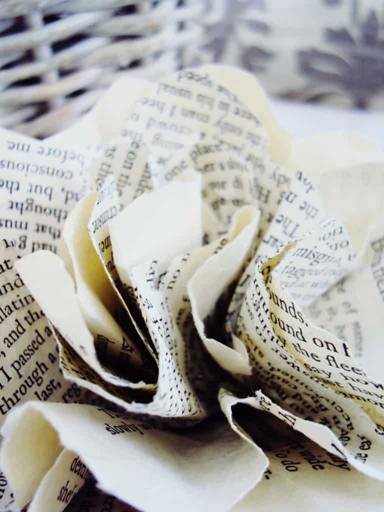 A close-up of an elegant paper flower crafted from book pages, with a blurred basket in the background hinting at a cozy, literary-themed decor.