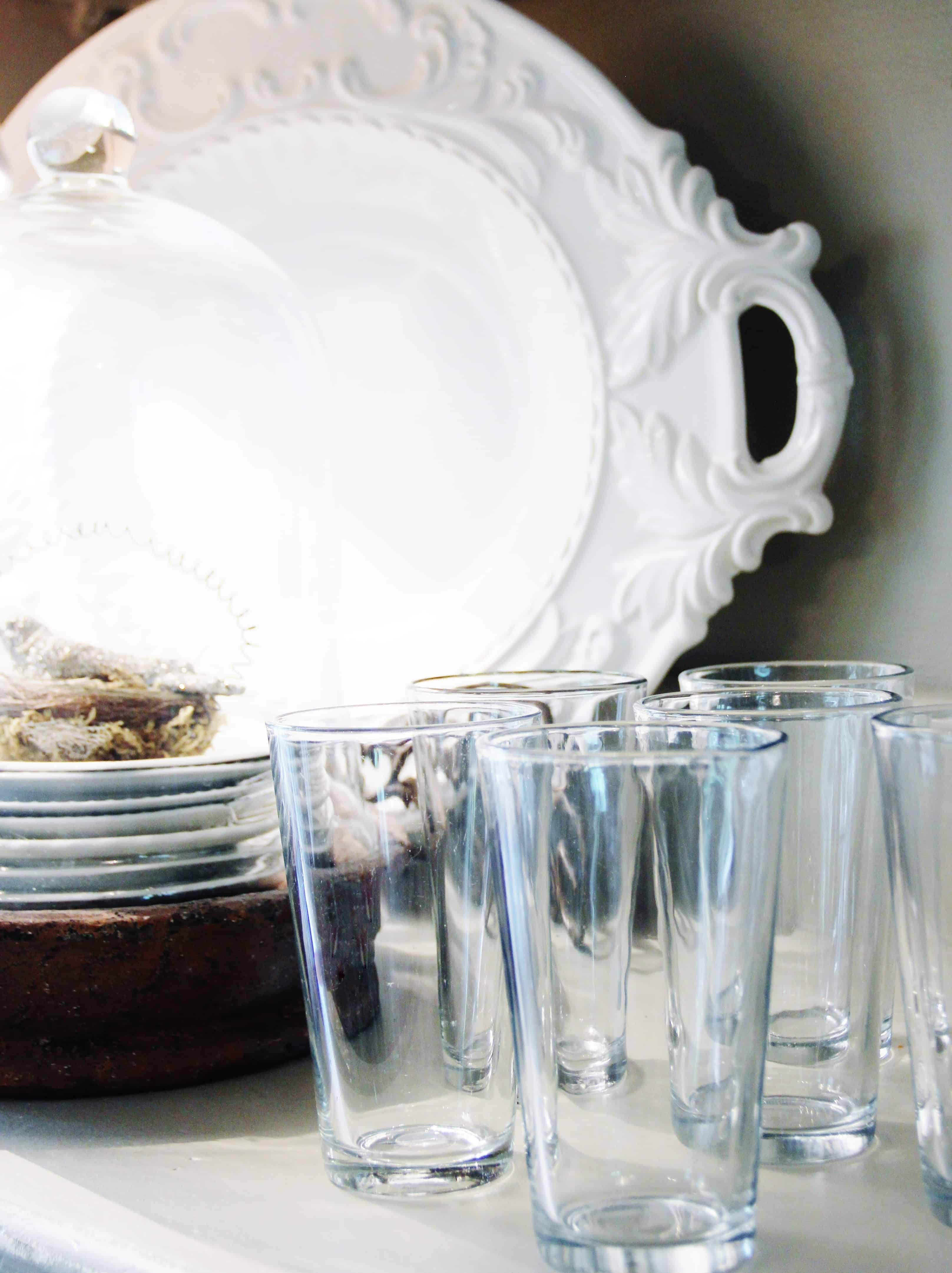 A collection of DIY etched glass tumblers neatly aligned in front of an elegant white decorative plate on a shelf, creating a cozy and organized kitchen vignette.