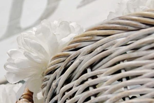Close-up of two woven wicker baskets with a white artificial flower tucked behind them. The background features blurred, large pale letters on a light surface.