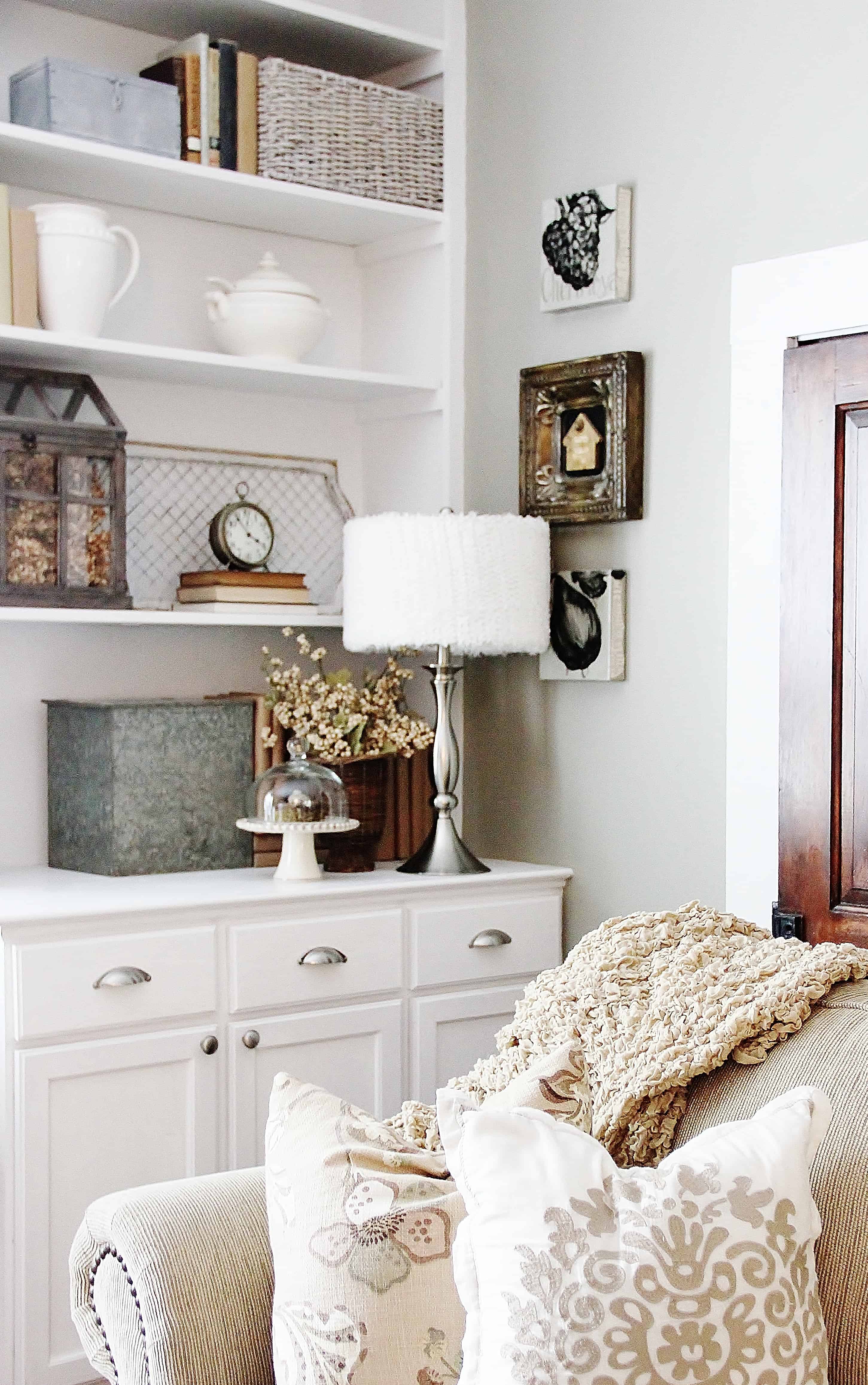 A cozy and tastefully arranged living space featuring an elegant white cabinet with a variety of decorative items on open shelves, a comfortable sofa with patterned cushions, and subtle touches of rustic charm.