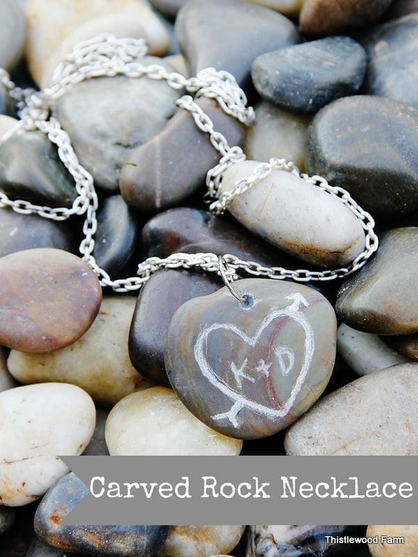 A heart and initial carved on a smooth stone pendant, part of a unique rock necklace, resting on a bed of assorted pebbles.