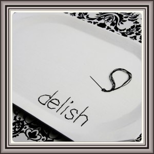 Image (5) An elegantly presented white serving dish with the word 'delish' playfully scribbled on its surface, accompanied by a large, decorative safety pin, all set against a striking black and white patterned backdrop.