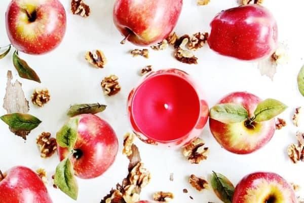 Flat lay of fresh red apples, a red candle, walnuts, and leaves scattered against a white background.
