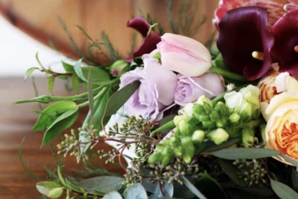A close-up of a bouquet featuring pink, purple, maroon, and cream flowers with green foliage, resting on a wooden surface with a blurred wooden background.