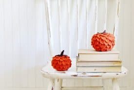 A vintage white chair with peeling paint against a white panelled wall, adorned with a stack of books and two decorative felted wool pumpkins in shades of orange and peach.