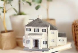 A small model of a white house with a dark roof sits on a closed book, surrounded by potted green plants and soft natural light—a charming scene perfect for summer decorating ideas in a cozy indoor setting.