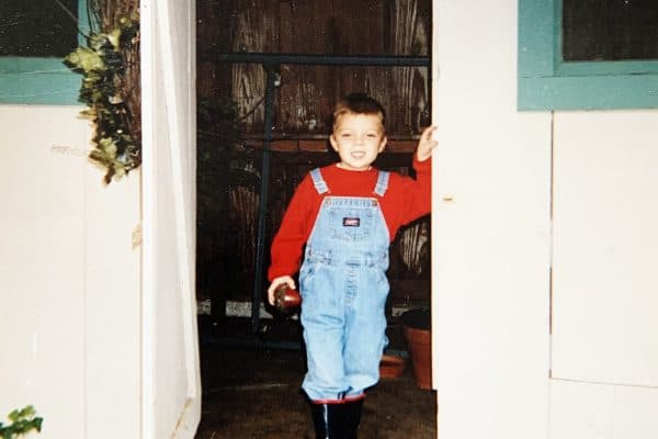 A young child smiles broadly while wearing blue overalls and standing in an open doorway framed by white and teal trim.