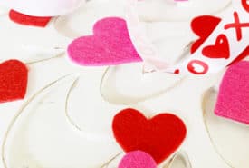 An assortment of pink and red felt hearts scattered on a white background, intertwined with a decorative ribbon featuring "xoxo" and heart patterns.