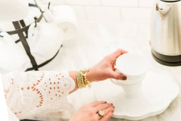A person wearing a white lace blouse lifting a white coffee cup from a saucer on a kitchen counter, with a kettle and cups in the background.
