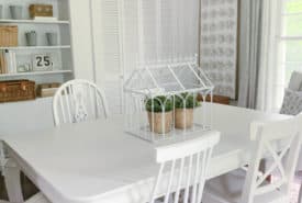 A bright dining room with a white table and chairs, featuring a decorative birdcage centerpiece with green plants on the table. bookshelves and patterned curtains complement the serene decor.