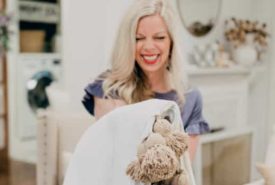 A smiling woman with blonde hair, wearing a blue dress, holding a plush toy covered by a white blanket in a cozy living room setting.