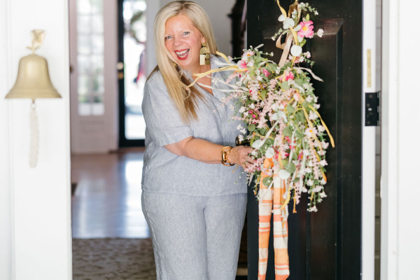 A smiling woman stands at an open front door, holding a large, colorful floral wreath decorated with ribbons. She is wearing a light blue outfit and gold sandals, welcoming someone into a bright, inviting home.
