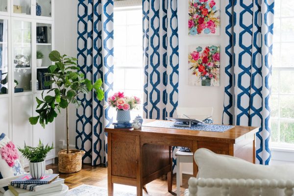 Bright home office with a wooden desk, patterned blue and white curtains, floral artwork, a large leafy plant, white bookshelf, and a cozy chair with a textured frame on a blue-patterned rug.