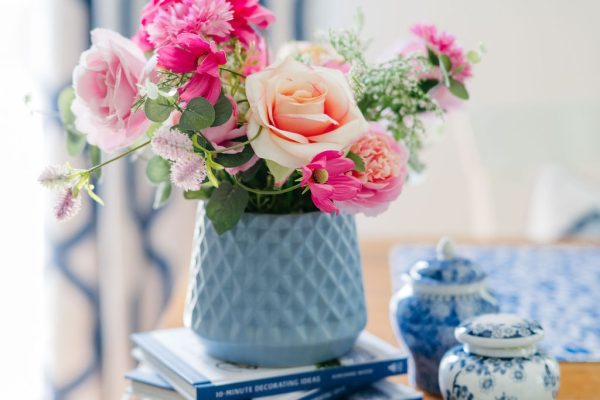 A blue vase with pink and peach flowers sits on stacked books atop a wooden table, next to two small blue-and-white ceramic jars. A blue patterned curtain is visible in the background.