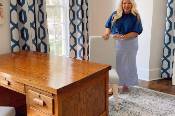 A woman with long blonde hair stands smiling beside a white chair in a bright room with a wooden desk, patterned rug, and blue-and-white curtains, showcasing one of the best dining chair slipcovers that adds both elegance and comfort to the space.