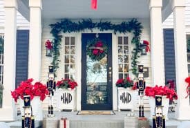 Festive house exterior inspired by Christmas decorating ideas for the porch, featuring red bows, garlands, and toy soldier statues flanking the entrance. Porch steps are adorned with gifts and poinsettias, while "Seven Hundred One" graces the door above in elegant display.