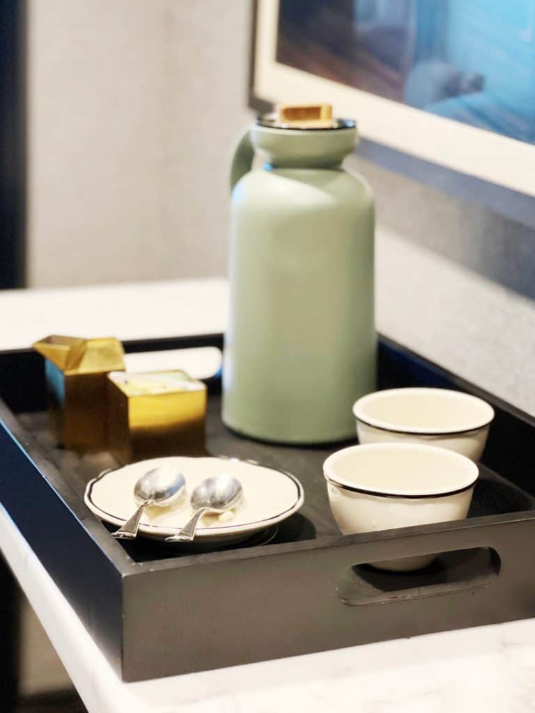 A modern tea set on a black tray including a mint green thermos, two white cups, and two spoons, elegantly arranged in a contemporary hotel room.