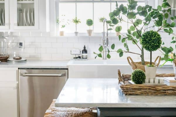 A bright, welcoming kitchen featuring white subway tiles, stainless steel appliances, and a contrasting island with gray cabinetry and a marble countertop, adorned with a woven basket and green plants, illuminated by natural light from the window.