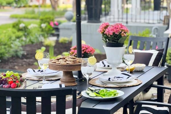 An outdoor dining table set for two, with plates of food, glasses of lemonade, and a vase of flowers, in a front yard setting with lush greenery in the background.
