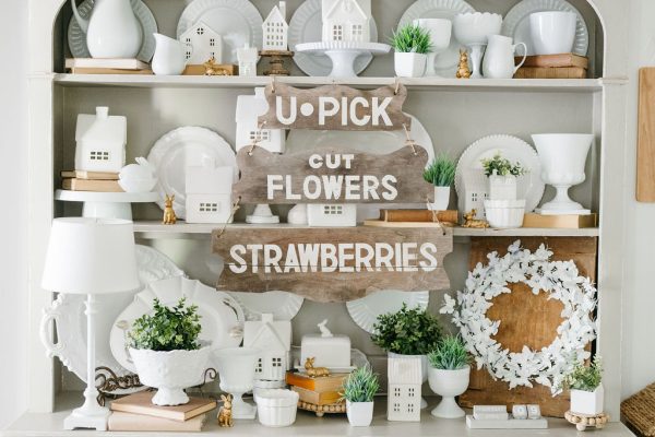 A beige hutch displays white dishes, pitchers, potted plants, and decorative items—showing how to save money by using thrifted finds. Three wooden signs read "U-PICK," "CUT FLOWERS," and "STRAWBERRIES." Wicker baskets sit on the top shelf.