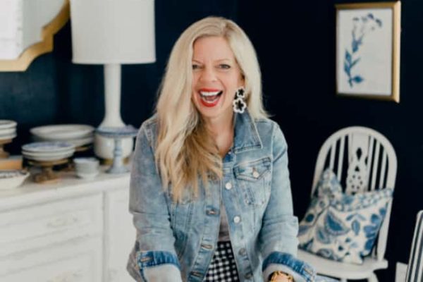 A smiling woman with long blonde hair, wearing a denim jacket and checkered skirt, stands at a table setting blue and white plates in a stylish dining room with dark walls and framed art.