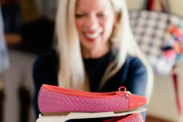 A smiling woman with long blonde hair is holding a pair of pink casual shoes. The shoes have a diamond pattern detail and orange tips, and are displayed on a white shoe stand. She is wearing a dark top with white stripes at the sleeves and hem.