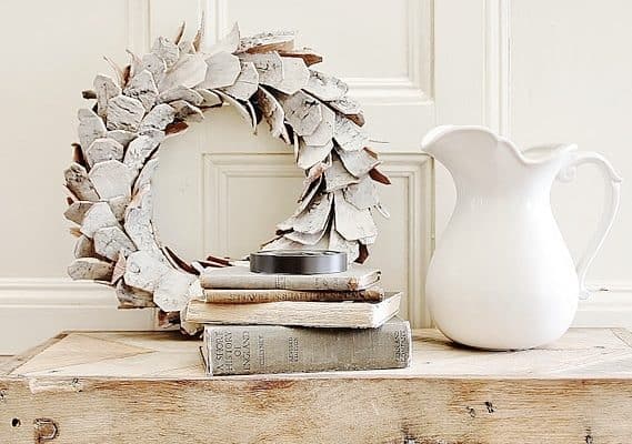 A white ceramic pitcher and a stack of vintage books sit on a rustic wooden table. A white and brown decorative wreath is propped up behind them against a paneled wall, creating a cozy, farmhouse-style scene.