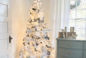 A white Christmas tree adorned with silver ornaments and snowflakes stands in a sunlit corner. It has a fluffy white skirt and a sign that reads "For to us a Savior is born." Beside it is a green dresser with nativity figurines.