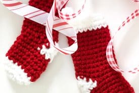 Two red and white crocheted Christmas stockings with decorative cuffs and toes are laid out on a white background. They are intertwined with red and white checkered ribbon, adding a festive touch.