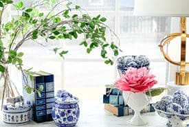 A white wooden table under a window, decorated with blue and white porcelain jars, a stack of books, and a decorative pink flower. A lamp and leafy branches add to the arrangement, with a blurred outdoor view in the background.