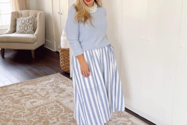 A woman in a light blue sweater and a blue-striped skirt smiles while standing on a patterned rug. The room features a white cabinet, a beige armchair, and woven baskets on top of the cabinet, with light streaming in through a window.