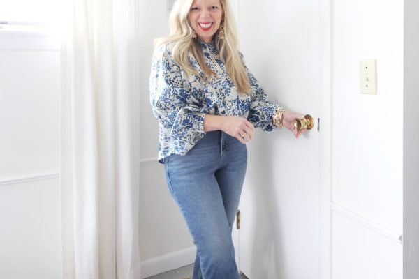 A person, smiling with enthusiasm for organizing a small closet, opens a white door. They're stylishly dressed in a blue patterned blouse, blue jeans, and chic white shoes with bows. The airy room has pristine white walls, a curtained window, and a cozy beige rug underfoot.