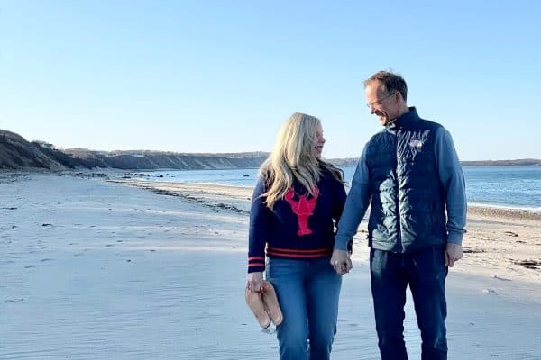 A couple walks hand in hand on a sandy beach. The woman is in a black sweater with a red design, holding sandals. The man wears a navy vest and jeans. The sky is clear and the ocean is visible to the right.