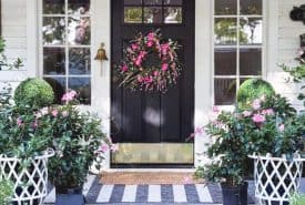 A black front door with a pink floral wreath is centered between two white lattice planters with greenery and flowers. Striped rugs and blooming plants decorate the steps, creating a welcoming porch.