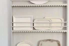 A white wooden shelf displays seven vintage ceramic plates of various shapes and sizes, including round, oval, and rectangular, all in neutral tones with some featuring subtle patterns or scalloped edges.