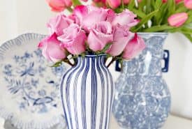 A blue and white striped vase filled with pink roses sits in front of a plate with a blue floral design and a blue and white patterned vase holding pink tulips.