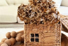 A woven basket shaped like a house with a small window holds dried brown hydrangeas. It sits on a wooden tray next to a decorative strand of large wooden beads, with a cream sofa in the background.