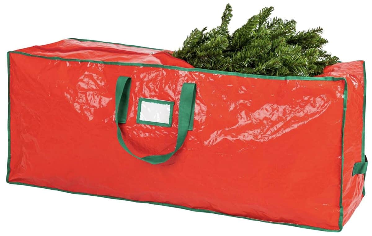 A large red storage bag with green handles and trim, partially unzipped to reveal artificial pine branches inside, likely for storing a Christmas tree.