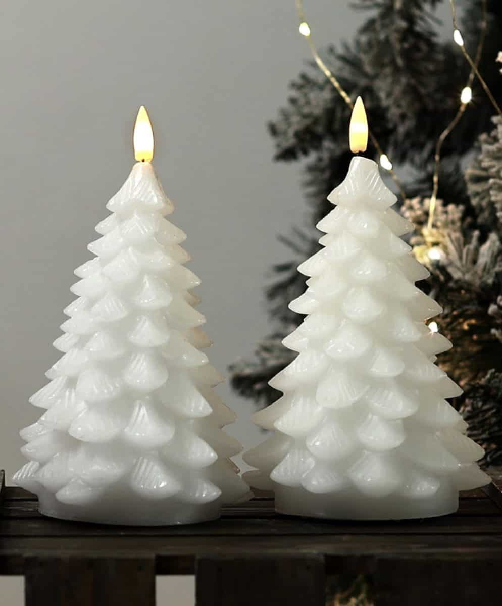 Two white candles shaped like Christmas trees are lit and placed side by side on a wooden surface, with a decorated tree and string lights in the background.
