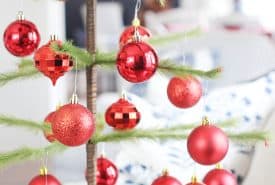 A minimalist Christmas tree with sparse green branches decorated with shiny and glittery red ornaments stands on a glass table in a bright, modern living room.