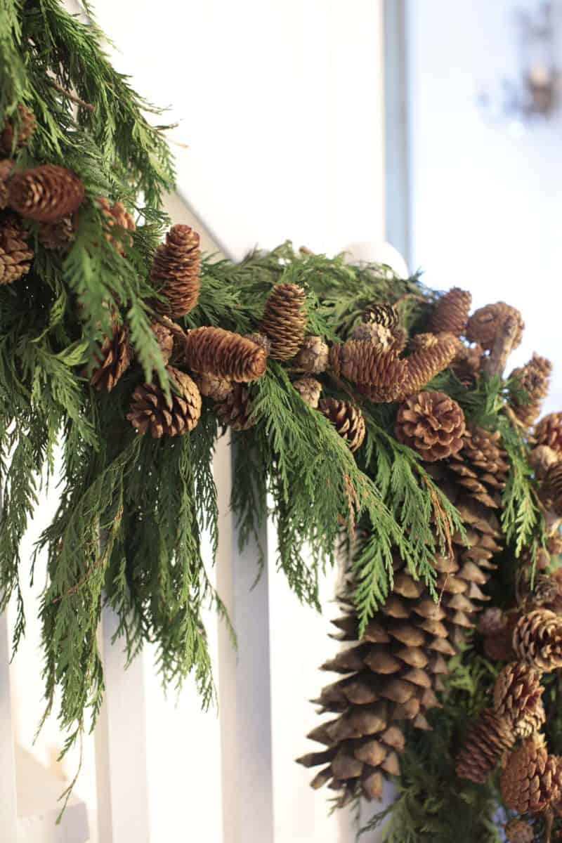 A garland made of green pine branches and brown pinecones is draped over a white stair railing, creating a festive, natural holiday decoration.